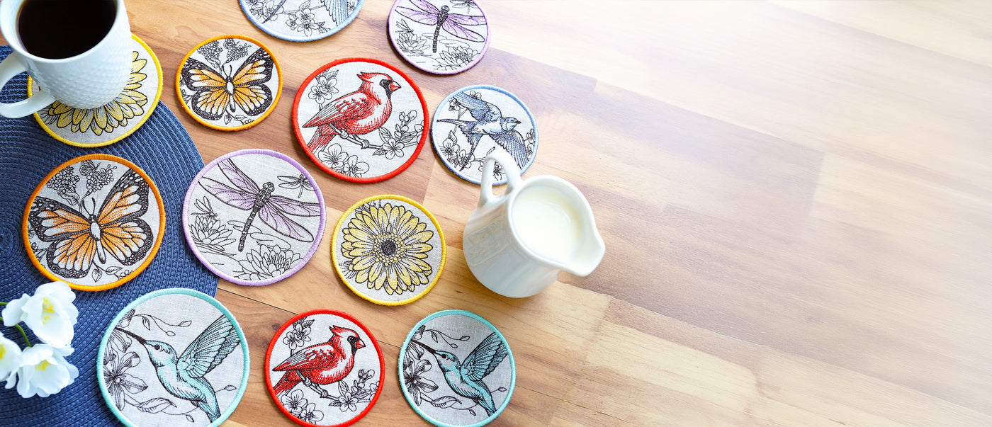 Round embroidered coasters featuring butterflies, birds, dragonflies, and floral designs arranged on a wooden table beside a coffee cup and creamer pitcher.