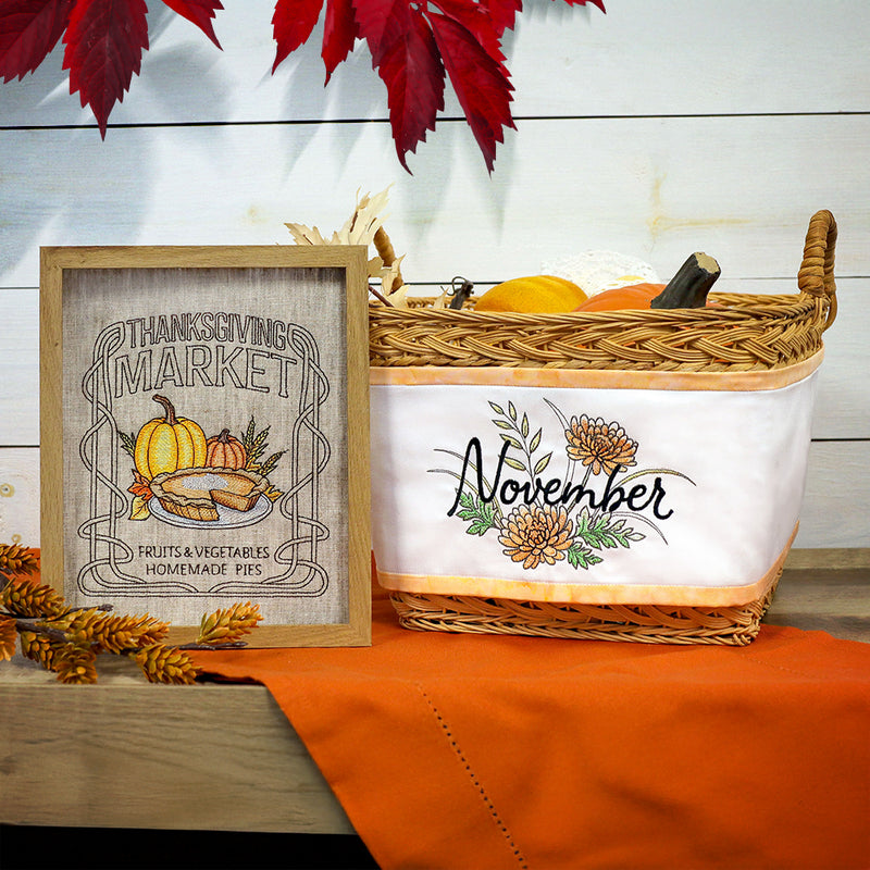 Embroidered Thanksgiving Market sign with pumpkins and pie next to a wicker basket labeled November with flowers, on an orange cloth and wooden table.