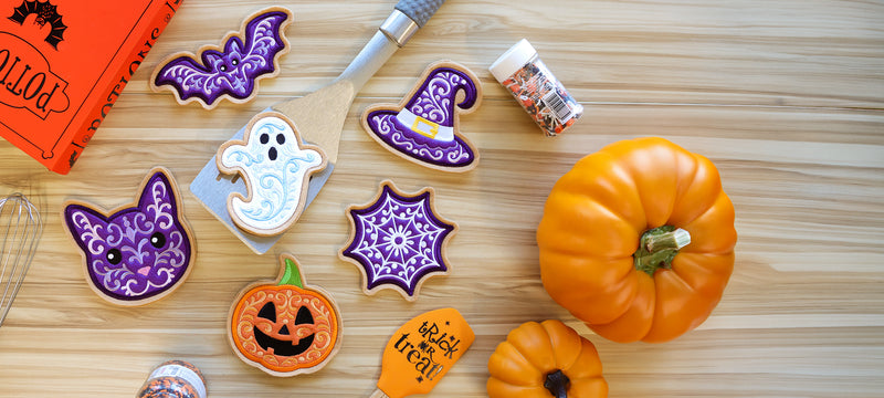 Halloween-themed embroidered cookies shaped like a bat, witch hat, ghost, black cat, spiderweb, and jack-o’-lantern arranged on a wooden surface with pumpkins and baking supplies.