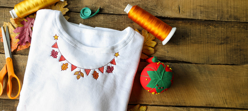 A folded white t-shirt with a colorful autumn-themed machine embroidered flag banner design on the chest, displayed on a rustic wooden table alongside sewing and crafting props including orange spools of thread, scissors, a pincushion, and leaves.