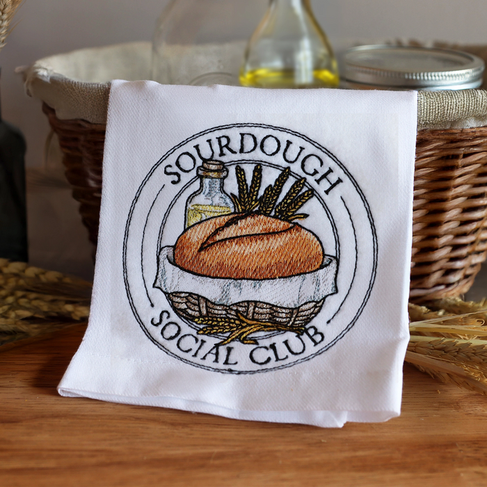 White towel with machine embroidery 'Sourdough Social Club' design on a wooden surface with wicker basket and bottles in the background.