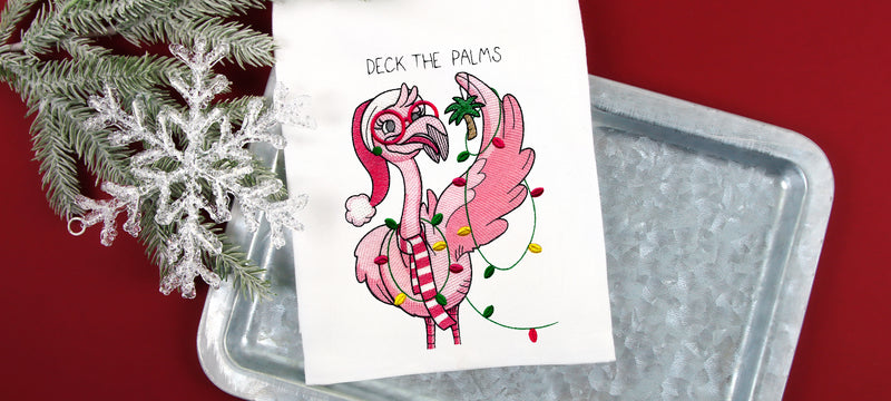 White towel with embroidered pink flamingo wearing a Santa hat and scarf, wrapped in colorful Christmas lights with the text "Deck the Palms" above.