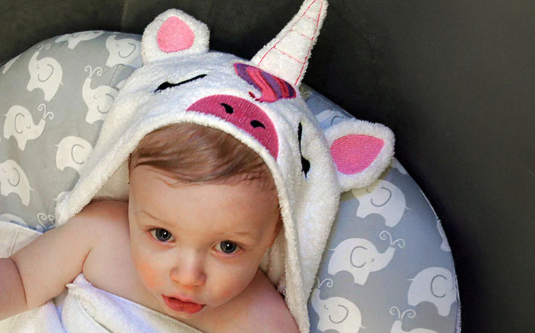 Baby wearing a unicorn-themed machine embroidered hooded towel sitting in a baby lounger.