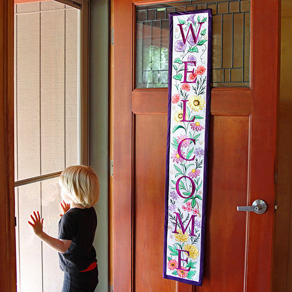 Child looking through a glass door with a decorative machine embroidered 'Welcome' banner on a wooden door.