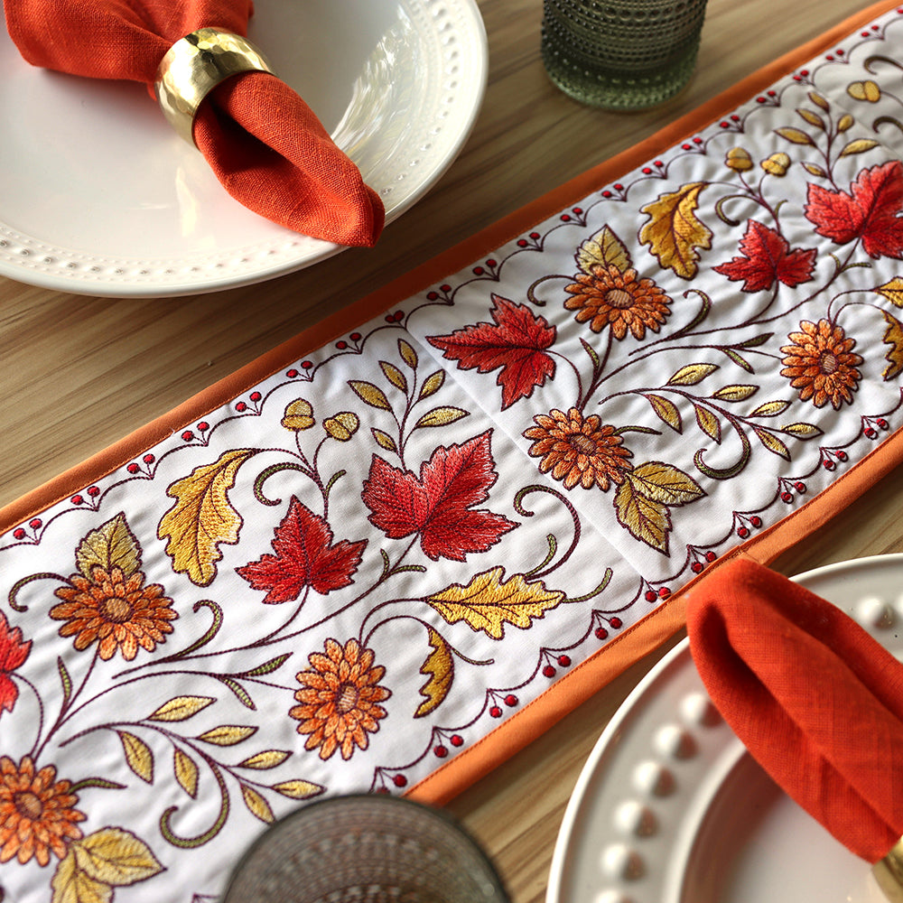 Machine embroidered autumn table runner with red, gold, and orange leaves and flowers, styled with orange napkins.