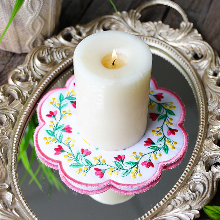 A lit white pillar candle sits centered on a decorative scalloped white fabric coaster with pink, green, and yellow floral machine embroidery, placed on an ornate silver mirror tray.
