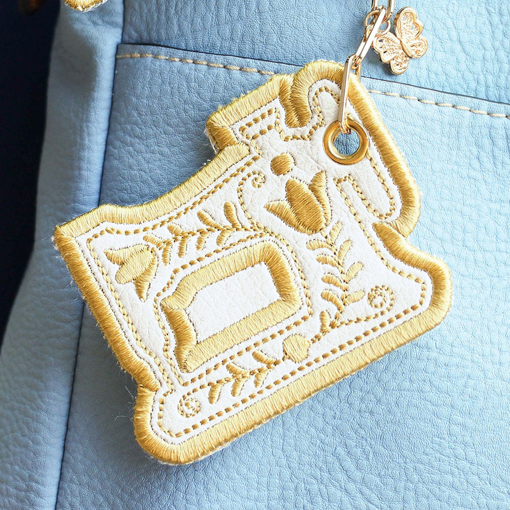 Cream and gold sewing machine embroidery with vintage details, shown as a finished keychain on a blue textured bag charm.