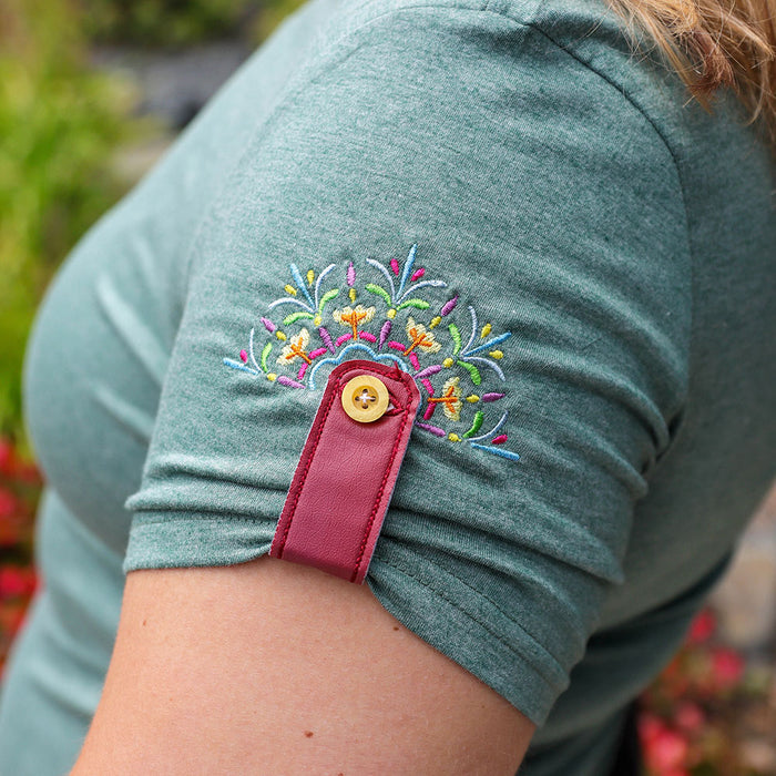 Close-up of a person's upper arm wearing a heathered sage green short-sleeve top with colorful floral embroidery on the shoulder and a magenta fabric cuff with a gold button.