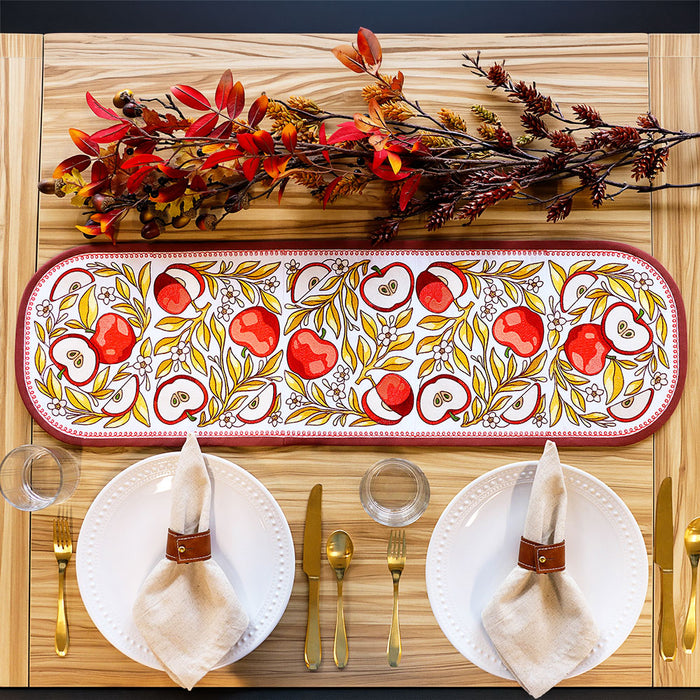 An autumn-themed machine embroidered table runner with a red and gold apple pattern, styled on a light wood table with plates, napkins, gold cutlery, and fall foliage.