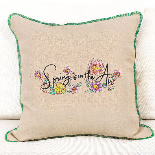 Decorative pillow with machine embroidery 'Spring is in the Air' text and floral design on a beige background.