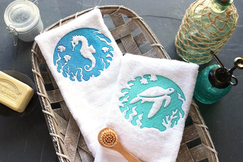 Two towels with machine embroidered embossed sea-themed designs on a tray with a bottle and soap.