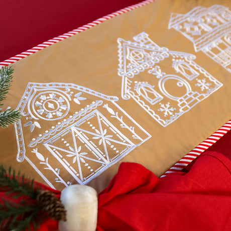 A beige fabric runner with white embroidered gingerbread-style houses is bordered by red and white striped edges, with red fabric and pine branches nearby.