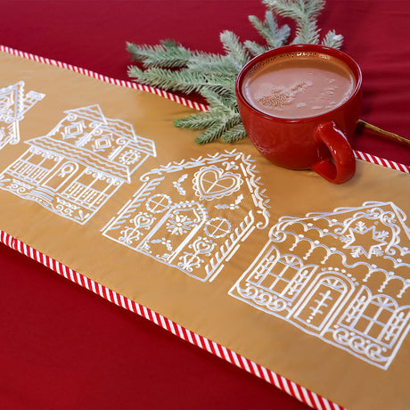 A red mug filled with hot cocoa sits on a tan table runner embroidered with white gingerbread house designs and a candy cane striped border, with a sprig of pine.