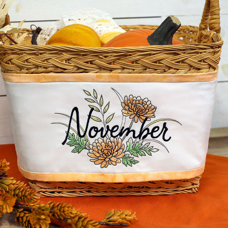 Wicker basket with white fabric side panel embroidered with "November" in black script and decorated with orange flowers and green leaves, filled with pumpkins.