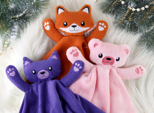 Three machine embroidered blanket buddies shaped like a fox, cat, and bear on a white surface with Christmas decorations.