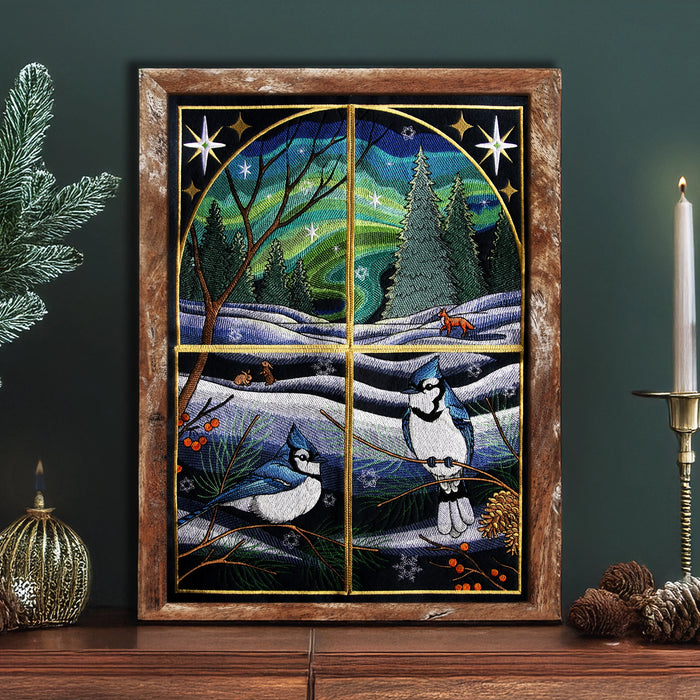 Framed machine embroidered design of blue jays in a winter landscape with stars and trees resting on a wooden table