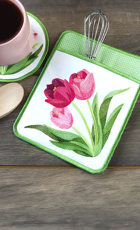 Machine embroidery floral pot holder with tulips on a wooden surface with a cup and utensils.