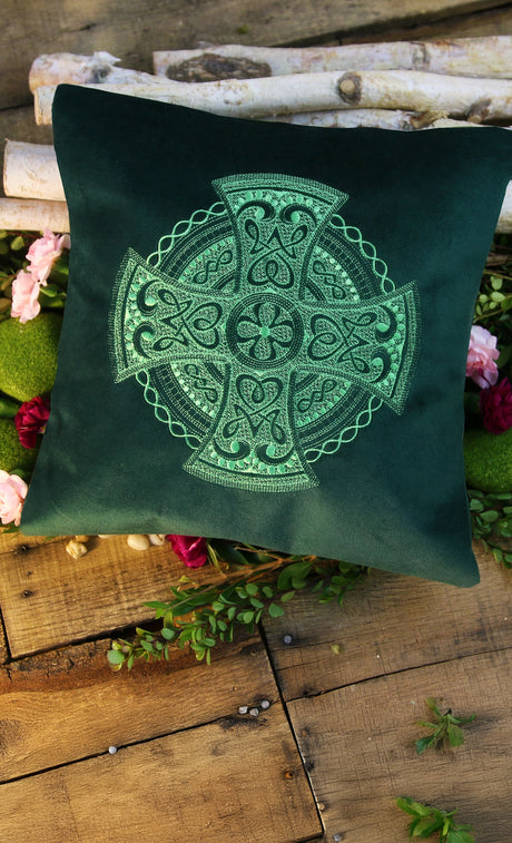 Green machine embroidered pillow with Celtic cross design on a wooden surface with plants.