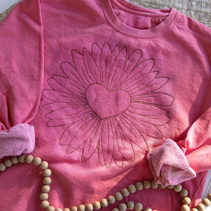 Pink sweatshirt with a machine embroidery heart and sunflower design on a textured surface.