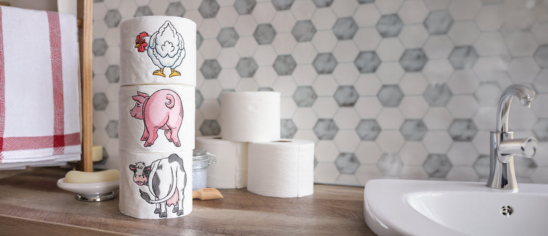 A stack of novelty machine embroidered toilet paper rolls decorated with farm animal embroidery designs (a chicken, a pig, and a cow) on a bathroom counter next to a sink and hexagonal tile backsplash.