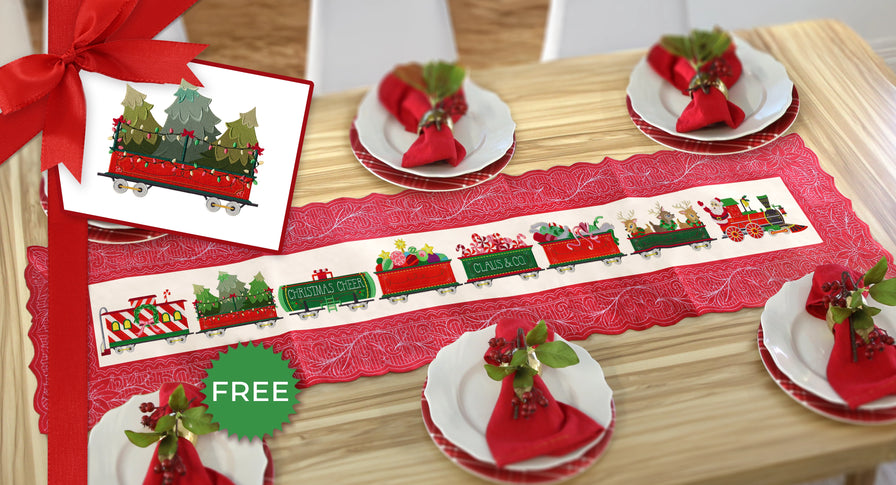 Christmas-themed table setting with machine embroidery train on a red tablecloth, greenery, and decorative elements on a wooden table.