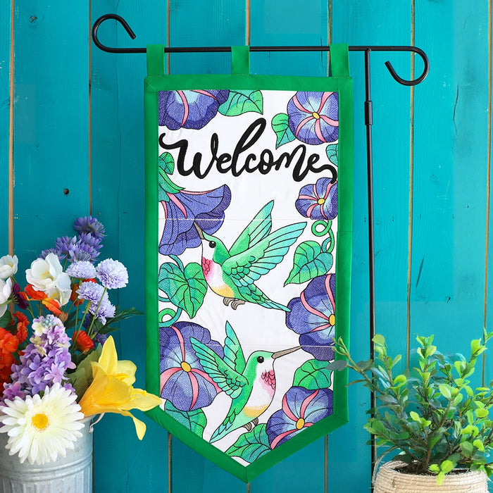 Decorative machine embroidery garden flag with hummingbirds and flowers against a teal wooden background.