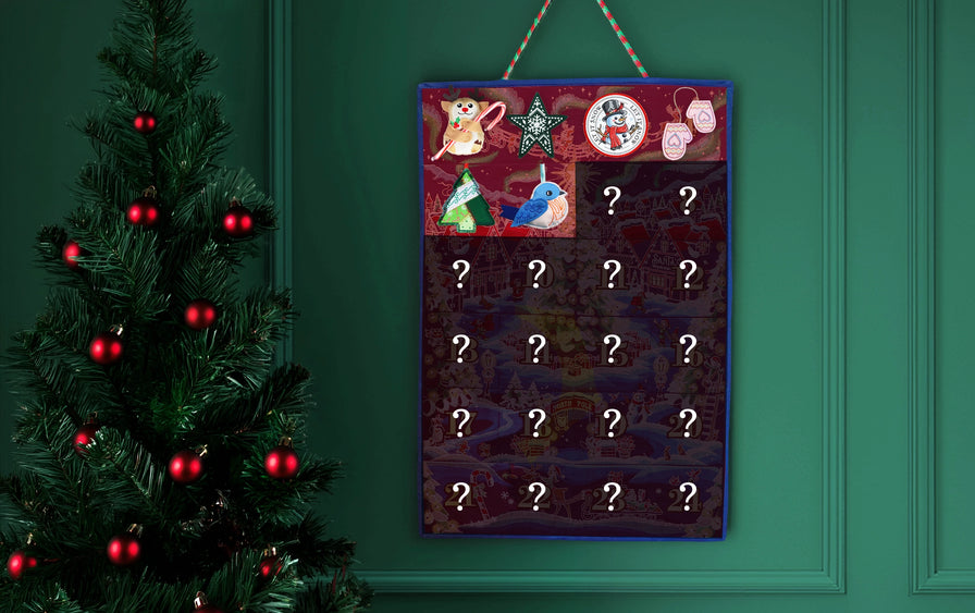 A Christmas advent calendar hanging on a dark green paneled wall next to a decorated Christmas tree. The calendar has colorful festive illustrations at the top and 20 numbered compartments with white question marks, suggesting hidden gifts.
