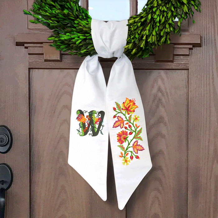 White fabric bow with colorful embroidered letter "W" and floral design in orange, red, yellow, and green, hanging on a green wreath on a brown door.