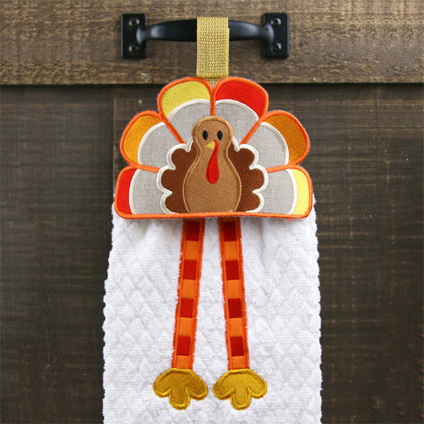 A turkey-themed embroidery design with colorful feathers is attached to a cabinet handle, showcasing a finished towel topper.