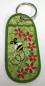 Green keychain with embroidered pink flowers and bee, green stitched outline, silver key ring attached.