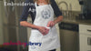 Video of someone wearing a white apron with a machine embroidery cupcake design and text 'embroidering on aprons'